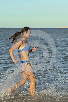 A sexy young brunette woman or girl wearing a bikini running through the surf on a deserted tropical beach with a blue