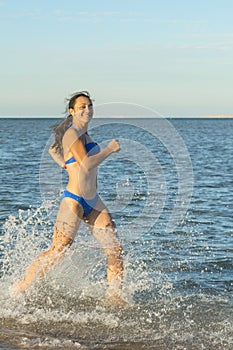 A sexy young brunette woman or girl wearing a bikini running through the surf on a deserted tropical beach with a blue sky. Young