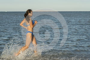 A sexy young brunette woman or girl wearing a bikini running through the surf on a deserted tropical beach with a blue sky. Young