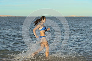 A sexy young brunette woman or girl wearing a bikini running through the surf on a deserted tropical beach with a blue sky. Young