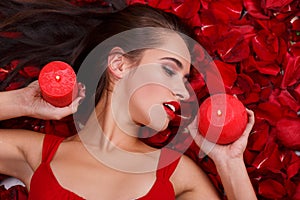 A girl lies in the rose petals, holds two burning candles and looks away.