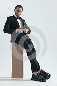 sexy young groom with glasses sitting and unbuttoning black tuxedo