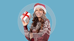 Sexy young lady in Santa hat and warm sweater holding Christmas gift on blue studio background, panorama