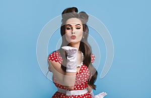 Sexy young pinup woman in old fashioned dress and gloves blowing air kiss at camera over blue background