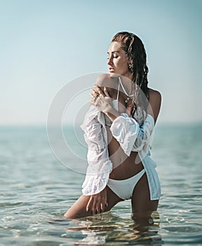 Sexy young slim woman girl in white bikini and shirt stands in sea touching her shoulder feeling softness. Summer vibes