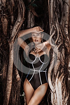 Sexy young woman in black bodysuit swimsuit stands in branches of tropical tree, lianas. Summer vibes photo