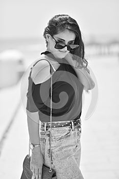 Sexy young woman on black t-shirt, fashion sunglasses and blue jeans with red fashion bag, stand on the beach. Girl doing sunbath