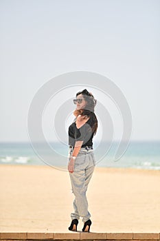 Sexy young woman on black t-shirt, fashion sunglasses and blue jeans stand on the beach, looking at the sea. Girl doing sunbath on