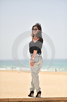 Sexy young woman on black t-shirt, fashion sunglasses and blue jeans stand on the beach, looking at the sea. Girl doing sunbath on