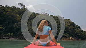 Sexy young woman in blue swimsuit swings feet in water on pink canoe on sea against hills backside view.