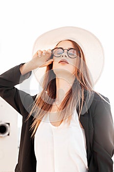 Sexy young woman in fashionable hat in stylish black jacket in white T-shirt straightens vintage glasses on bright sunny day