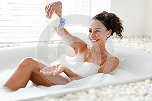 Sexy young woman lying in bubble bath near window, relaxing, adding foam into water, taking care of her body at home