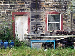 A shabby abandoned rural house with dirty windows and peeling paint overgrown with weeds with scrap and junk piled