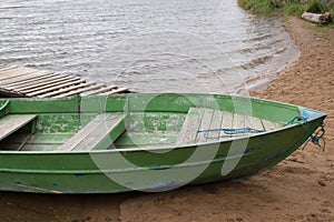 Shabby wooden boat painted green with two benches for rowing moored by rope at small board pier on shore of a bay of lake