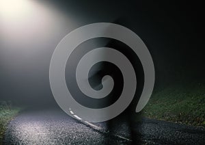 A shadow ghost like figure, on an empty spooky road at night. With a single street light in the countryside