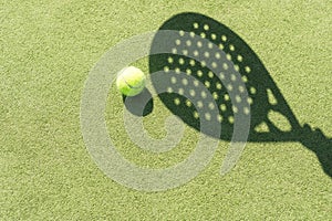 Shadow of a padel racket with a yellow ball on the green grass.