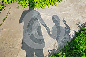 Shadows of mother with baby on cement pavement next to lawn in which son shows finger where to go and what to see