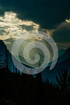 Shafts of light break through the stormy clouds onto the steep mountain valley slope, silhouetting trees in the foreground