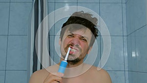 Shaggy bearded man brushing teeth using electric toothbrush in bathroom.