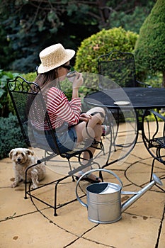 A shaggy dog sits on the terrace and a girl sits on a chair and drinks coffee from a cup.