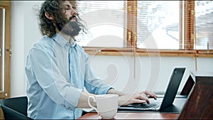 Shaggy and sleepy man at the desk with a laptop drinks from a cup of coffee and becomes workable and cheerful. The drink