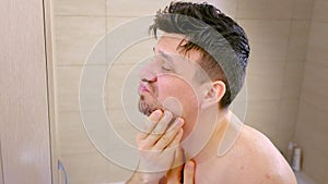 Shaggy unshaven man looks at himself in bathroom mirror in morning, side view.