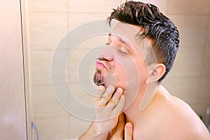 Shaggy unshaven man looks at himself in bathroom mirror in morning, side view.