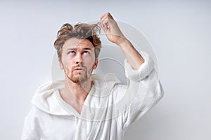 shaggy young guy in bathrobe isolated on white background, touches long regrown hair