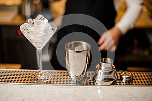 Shaker and cocktail glass filled with ice cubes on the bar counter