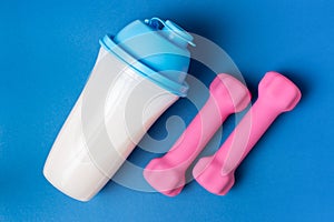 Shaker with protein and pink dumbbells on blue background, top view, sports nutrition concept