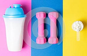 Shaker with protein, pink dumbbells, measuring spoon with protein powder, on pink blue yellow background, top view