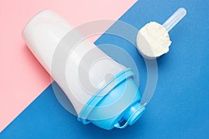 Shaker and scoop with protein on pink and blue background, top view.