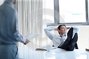 Shaking his responsibilities. A laidback businessman with his feet up on his desk as a coworker stands by.