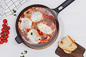 Shakshuka egg dish with tomato sauce in a black cast iron pan on the white wooden table