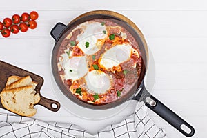 Shakshuka egg dish with tomato sauce in a black cast iron pan on the white wooden table