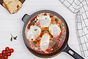 Shakshuka egg dish with tomato sauce in a black cast iron pan on the white wooden table