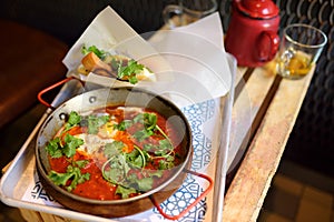 Shakshuka at pan on tray in cafe of Israeli cuisine. Shakshouka is fried eggs with tomatoes, vegetables and herbs