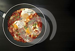 Shakshuka - poached eggs in tomato sauce, onion, pepper and spices in iron pan on dark wooden table with copy space