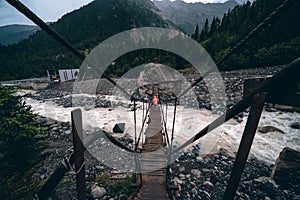 A shaky wooden bridge with metal cables. Stormy flow of a mountain river. Active recreation and hiking. Gloomy rainy weather.