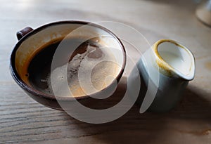 Shallow focus on black coffee in a rustic style pottery cup next to milk in a small jug. It is on a wooded table lit by window