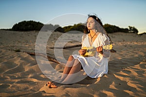 Shallow focus shot of a female sitting on a sandy ground while playing a yellow ukulele at the beach