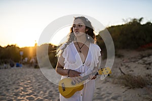 Shallow focus shot of a female in a white dress posing at the beach while holding a yellow ukulele