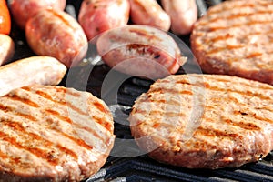 Shallow focus shot of tasty assorted delicious sausages and burgers sizzling and cooking on a barbecue griddle plate, outside in b