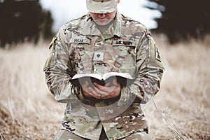 Shallow focus shot of a young soldier kneeling on a dry grass while reading the bible