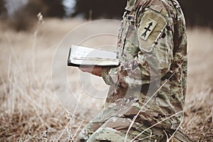 Shallow focus shot of a young soldier kneeling while holding an open bible in a field