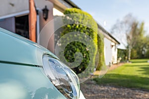 Shallow focus of a small cars headlight, seen parked in a private drive next to a garage.