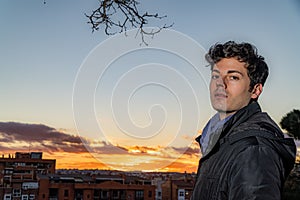 Shallow focus of a Spanish young man in a thick jacket against a beautiful sunset sky over a village