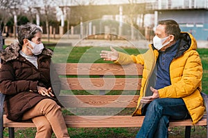 Shallow focus of two adult people with face masks sitting apart on a bench and chatting