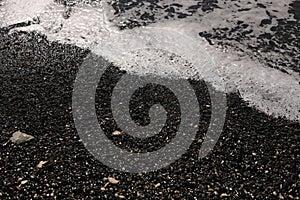 Shallow volcanic black sandy beach and pebbles, water waves and white foam washes the coastline with wet dark sand on the sea