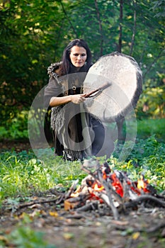 Shaman woman drumming next to the fire in the evening forest